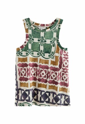 Femme portant un haut sans manches à motifs et un pantalon large crème, debout dans un salon beige avec un canapé et une plante.