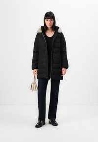 Cappotto trapuntato nero con cappuccio in pelliccia sintetica, zip anteriore e tasche laterali. Abbinato a jeans scuri e scarpe nere, mentre tiene in mano una piccola borsa beige.