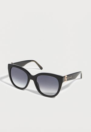 Czarne, dużych rozmiarów okulary przeciwsłoneczne w stylu kociego oka z gradientowymi szarymi soczewkami i złotym logo Marc Jacobs na zausznikach na białym tle.