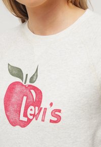 Bílá mikina s červeným motivem jablka a nápisem „Levi's“ v růžovém odstínu. Má kulatý výstřih a texturovanou látku.