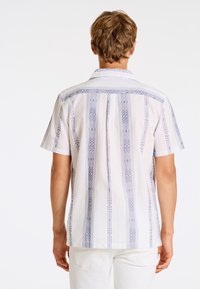 Camisa de manga corta blanca con patrones tribales azules; diseño con cuello, corte recto y una textura sutil.