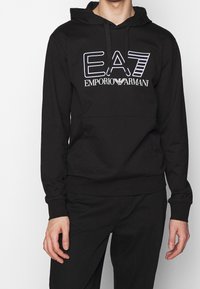 Felpa nera con cappuccio in misto cotone, dotata di tasca a marsupio frontale e logo EA7 bianco audace sul petto. Tessuto dalla texture liscia.