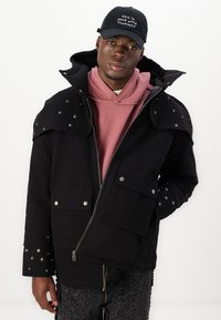 Chaqueta negra con cremallera frontal, gran capucha y acentos de tachuelas en los hombros. Combinada con una sudadera rosa y pantalones negros texturizados.