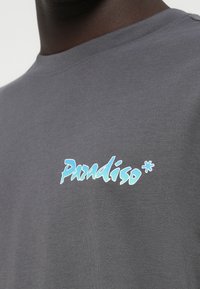 Graues Baumwoll-T-Shirt mit einem blauen stilisierten "Paradiso*"-Logo auf der Brust. Rundhalsausschnitt und kurze Ärmel mit einer glatten Textur.