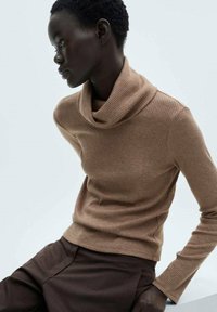 Pull en laine brun à col roulé avec une texture côtelée, présentant un décolleté cowl ample, associé à un pantalon noir marron foncé à jambes fuselées.