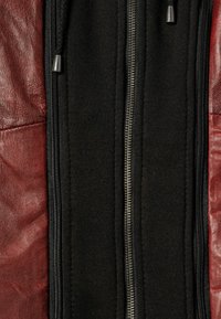 Leder- und Stoffjacke mit einem tiefroten Lederpanel und einem schwarzen Stoffmittelteil, ausgestattet mit einem silbernen Reißverschluss und schwarzen Kordelzügen.
