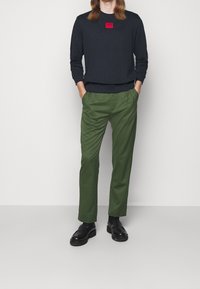 Felpa navy con patch logo rosso, abbinata a pantaloni tapered verdi e loafers neri, con una vestibilità comoda e tasche laterali.