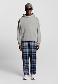 Uomo che indossa un hoodie grigio chiaro, un berretto di maglia verde, occhiali da sole scuri, pantaloni a scacchi blu e neri e sneaker bianche, in piedi contro uno sfondo grigio.