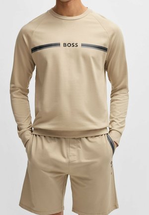 Mann trägt beige Langarm-Sweatshirt mit dem Schriftzug "BOSS" sowie passende Shorts mit Seitentaschen und schwarzen Streifendetails.