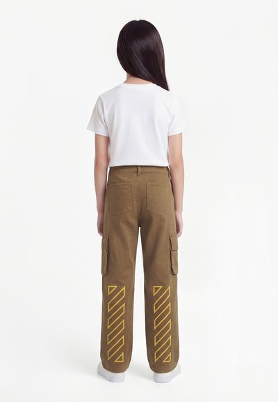 Παιδί που στέκεται με την πλάτη προς τα εμπρός, φοράει άσπρο μπλουζάκι και καφέ παντελόνι cargo με κίτρινες διαγώνιες ρίγες στα πίσω πόδια.