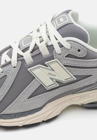 Grå sportig sko med ett texturerat mesh-ovandel, vita skosnören och en krämfärgad sula med en synlig dämpningsteknikdetalj.