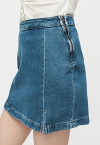 Blå denim mininederdel med synlige sømme, sidelukning med lynlås og lille frontslids, båret med en hvid top stoppet ind.