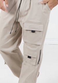 Beige Cargo-Hosen mit mehreren Taschen, Reißverschlüssen und schwarzen Hardware-Akzenten. Hergestellt aus leichtem Stoff mit lockerer Passform und verstellbarem Kordelzug.
