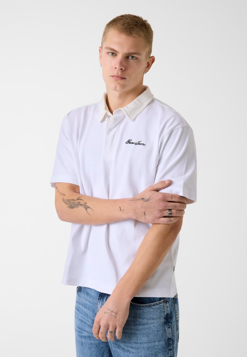 Giovane uomo con capelli biondi corti, indossa una polo bianca e jeans blu, braccia incrociate, mostra tatuaggi sugli avambracci e dita.