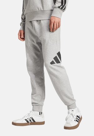 Uomo che indossa pantaloni della tuta Adidas grigi e felpa abbinata con strisce nere e sneaker bianche Adidas con suola in gomma.