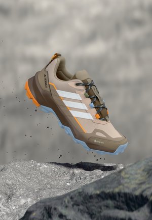 Light brown and orange hiking shoe with white stripes and blue sole, suspended above rocky surface with dirt particles falling.