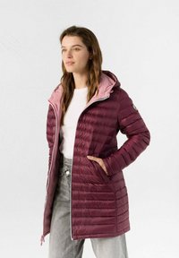Giacca imbottita color marrone bordeaux con cappuccio rosa chiaro, design trapuntato, chiusura con zip e tasche laterali. Indossata sopra una maglietta bianca e jeans grigi.