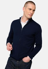 Cardigan zippé marine, en matière tricotée, col montant, poignets et ourlet côtelés. En dessous, une chemise blanche à col est partiellement visible.