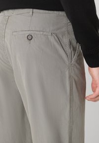 Grå bomuldsbukser med en glat tekstur, der har en baglomme med knappelukning og sidelommer. Syede detaljer langs sømmene.