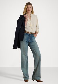 Cream ruffled blouse, blue wide-leg jeans, brown belt, and dark jacket. Model stands casually with hand on hip. Textured fabrics visible.