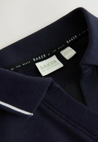 Polo azul marino con cuello de punto plano, textura fina y franja blanca decorativa. Etiquetas visibles: "BAKER by Ted Baker", fabricado en Pakistán.