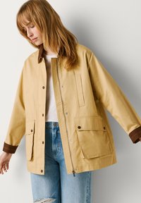 Sieviete valkā bēša krāsas žaketi ar brūnām aprocēm virs baltas krekla un gaiši zilas saplēstas džinsa bikses, stāvot pret vienkāršu gaišu fonu.