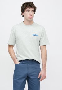 Γκρι βαμβακερό μπλουζάκι με κοντά μανίκια, με μπλε λογότυπο "DICKIES" στο αριστερό στήθος. Συνδυάζεται με μπλε παντελόνι, παρουσιάζοντας μια casual εφαρμογή.