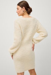 Vestito maglia color crema fuzzy con maniche lunghe, vestibilità rilassata e superficie testurizzata, caratterizzato da un collo rotondo e un orlo leggermente svasato.