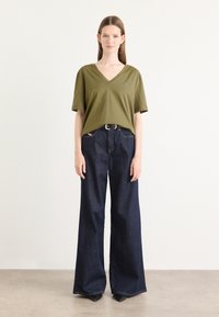 Olivově zelené tričko s výstřihem do V vyrobené z jemné tkaniny, doplněné širokými tmavými džíny. Outfit obsahuje úzký černý opasek a decentní logo.