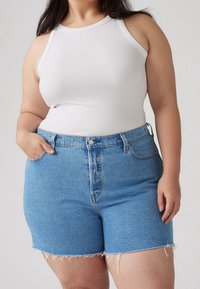 Débardeur blanc côtelé associé à un short en denim bleu clair taille haute, avec ourlet effiloché et fermeture à boutons sur le devant.