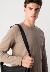 Beige lange mouw ribgebreide shirt met een ronde hals, gedragen door een persoon die een zwarte schoudertas met ritsdetail vasthoudt.