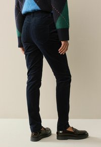 Marineblå corduroybukser med slim fit, der har to baglommer og er stylet med sorte, blanke loafers. Baggrunden er i en neutral farve.