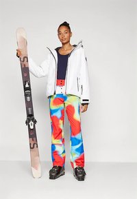 Lyžiarsky outfit zahŕňajúci bielu bundu, tmavomodrý top a farebné, víriace vzorované snehové nohavice. Modelka drží lyžu s viazaním.