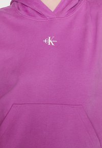 Purple hoodie featuring a front pocket, drawstring hood, and a white logo reading "Calvin Klein Jeans" on the chest. Smooth texture.