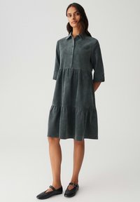 Vrouw staat in een donkergrijze, knoopsluiting, gelaagde midi-jurk met ellebooglange mouwen en zwarte Mary Jane flats, op een effen lichte achtergrond.
