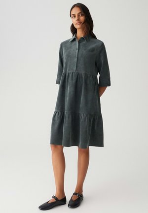 Femme debout portant une robe mi-longue à boutons de couleur gris foncé avec des manches mi-longues et des chaussures Mary Jane noires, sur un fond clair uni.