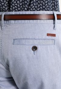 Jasnoniebieskie spodnie o gładkiej fakturze, z tylną kieszenią zapinaną na guzik i przeszyciami, do których dołączony jest brązowy skórzany pasek.