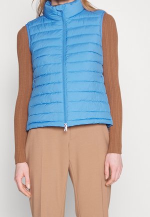Femme portant une veste matelassée sans manches bleu clair par-dessus un top marron à manches longues côtelé et un pantalon beige, debout bras détendus.
