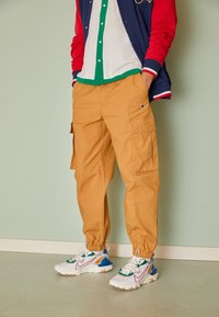 Gula cargobyxor med elastiska muddar, utrustade med sidofickor. I kombination med vita sneakers som har gröna, röda och blå detaljer.