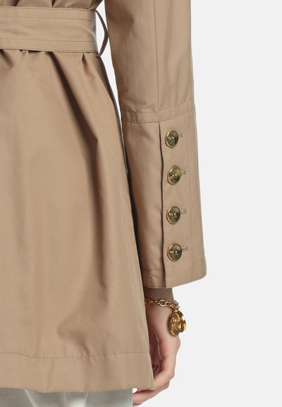 Beige trench coat detail featuring a belted waist, buttoned sleeve cuffs with gold buttons, and a smooth fabric texture.