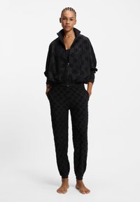 Sort afslapningssæt med jakke med lynlås og høj hals samt matchende bukser, begge med tekstureret logomønster og elastiske manchetter.