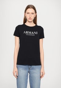 Svart bomull T-skjorte med "ARMANI EXCHANGE" logo i metallisk design. Korte ermer, rund hals og avslappet passform. Kombinert med lyseblå jeans.