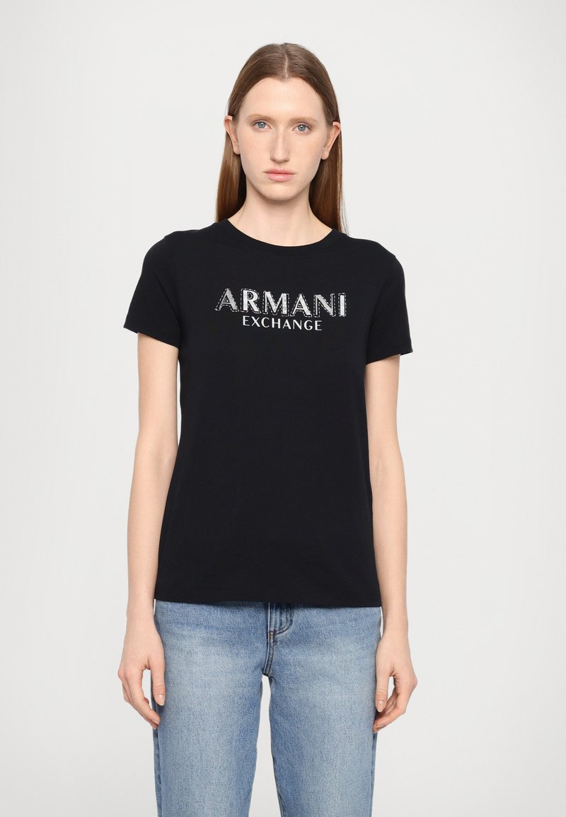 Svart bomull T-skjorte med "ARMANI EXCHANGE" logo i metallisk design. Korte ermer, rund hals og avslappet passform. Kombinert med lyseblå jeans.