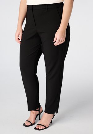 Vrouw met zwarte slim-fit enkellange broek en zwarte sandalen met hak, staand tegen een effen lichte achtergrond.