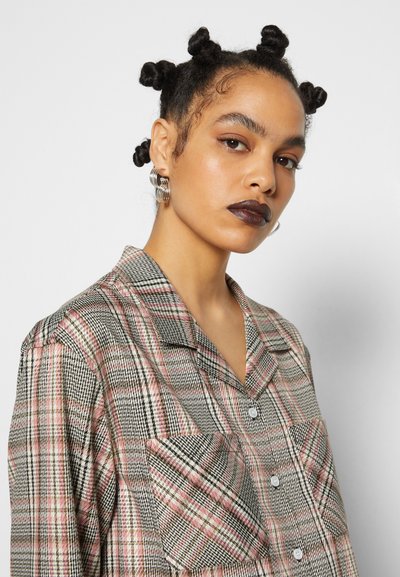 Camisa às listas com um padrão preto, branco e rosa, colarinho, fecho com botão e um bolso no peito. O modelo usa batom escuro e brincos.
