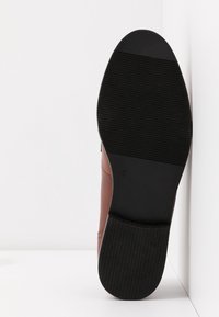 Semelle d'une chaussure en cuir marron avec un talon en caoutchouc noir et une semelle extérieure noire texturée, positionnée verticalement contre une surface blanche.