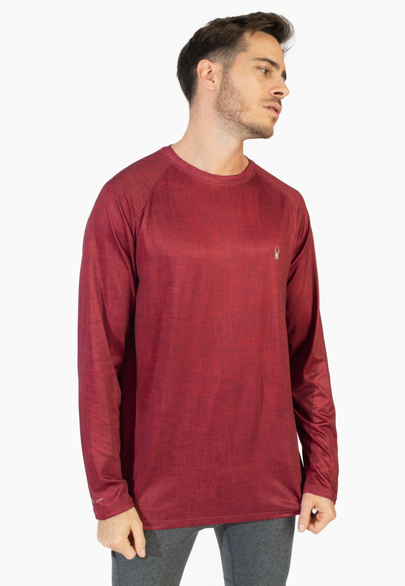 Mies, der ein langärmliges, strukturiertes rotes Hemd und graue Hosen trägt, schaut mit neutralem Ausdruck nach rechts vor einem weißen Hintergrund.