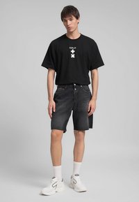 T-shirt nera con emblema bianco, abbinata a pantaloncini di denim scuro. Il modello indossa sneaker bianche e calzini alla caviglia, mostrando un look casual.