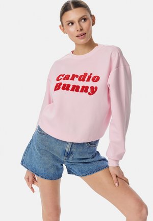 Kobieta ubrana w różową bluzę z czerwoną napisem „cardio Bunny” i jeansowe szorty, pozująca z jedną ręką na udzie i patrząca na bok.