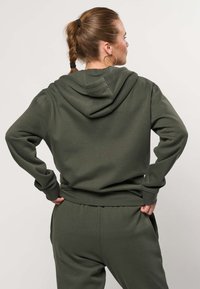 Frau mit geflochtenem Haar und Creolen, die rückwärts schaut, trägt einen dunkelgrünen Kapuzenpullover und passende Hosen, die Hände in die Hüften gestützt.